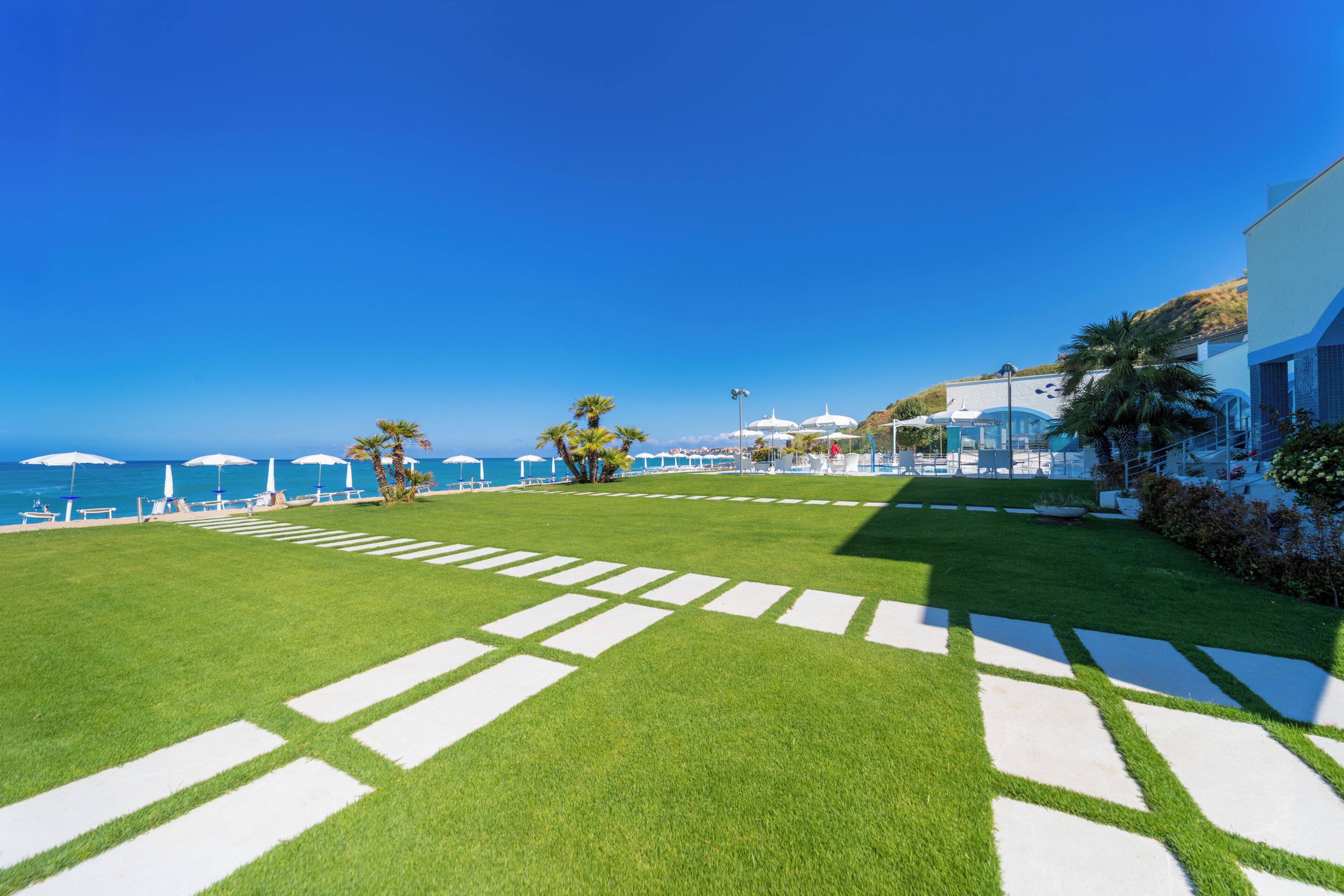 Une pelouse bien entretenue offrant une vue dégagée sur l’océan, un espace piscine agrémenté de parasols et un bâtiment au design moderne.