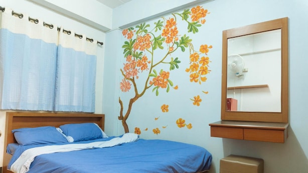 A bedroom with a bed, a nightstand, a mirror, and a wall with a tree and flowers mural. - Chiang Mai Tree Hostel (Chiang Mai)