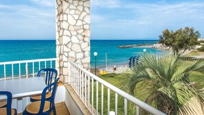A balcony with a stone pillar, a table, and blue chairs overlooking a beach with a clear blue sea and a rocky shoreline.