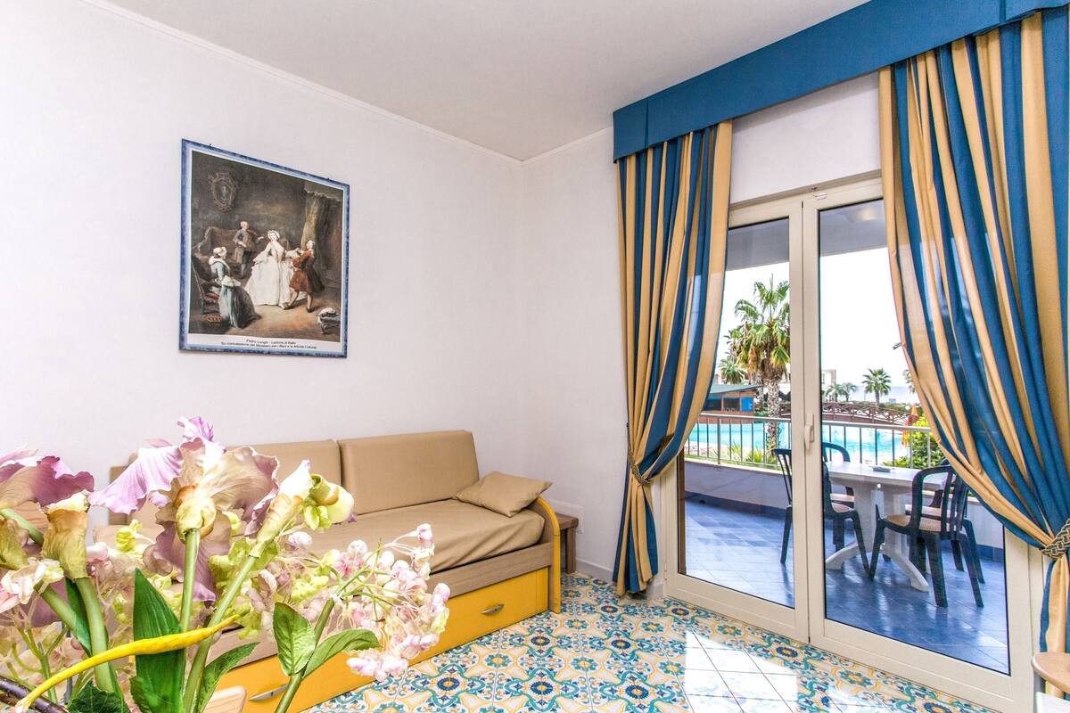 A room with a beige sofa, a large window with blue and yellow curtains, a tiled floor, and a framed painting on the wall.