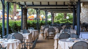 An outdoor dining area with tables and chairs, covered by a roof structure, and surrounded by greenery.