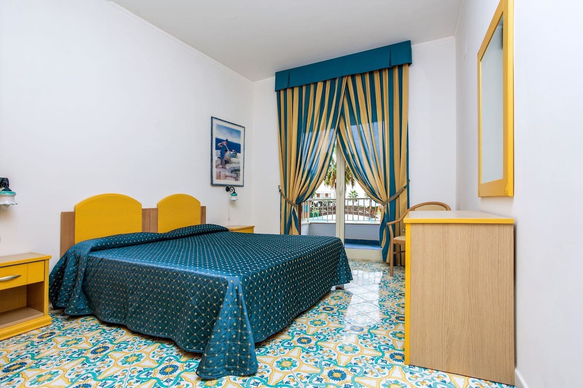 A bedroom with a bed, a wooden nightstand, a desk, and a window with curtains.