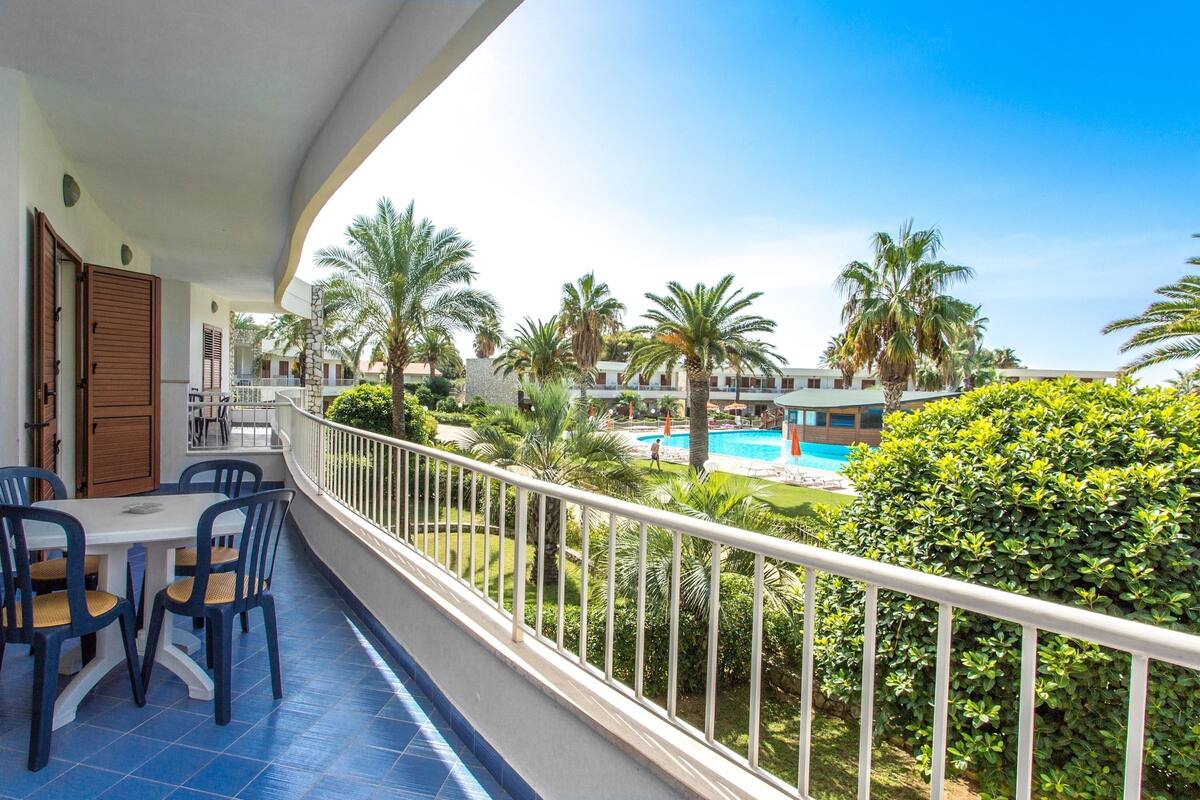 A balcony with a view of a swimming pool, palm trees, and a building in the background.