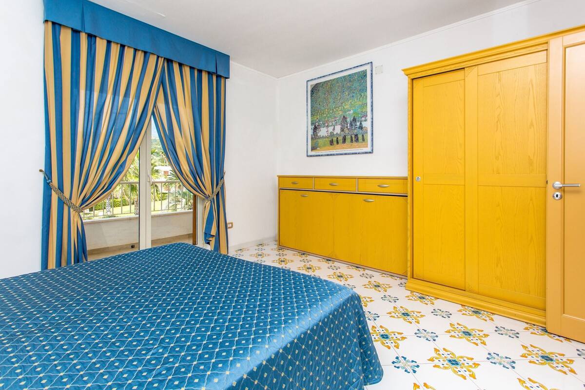 A bedroom with a bed, curtains, a yellow wardrobe, and a painting on the wall.