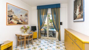 A room with a dining table, a chair, a small desk with a computer, a yellow cabinet, and a painting on the wall.