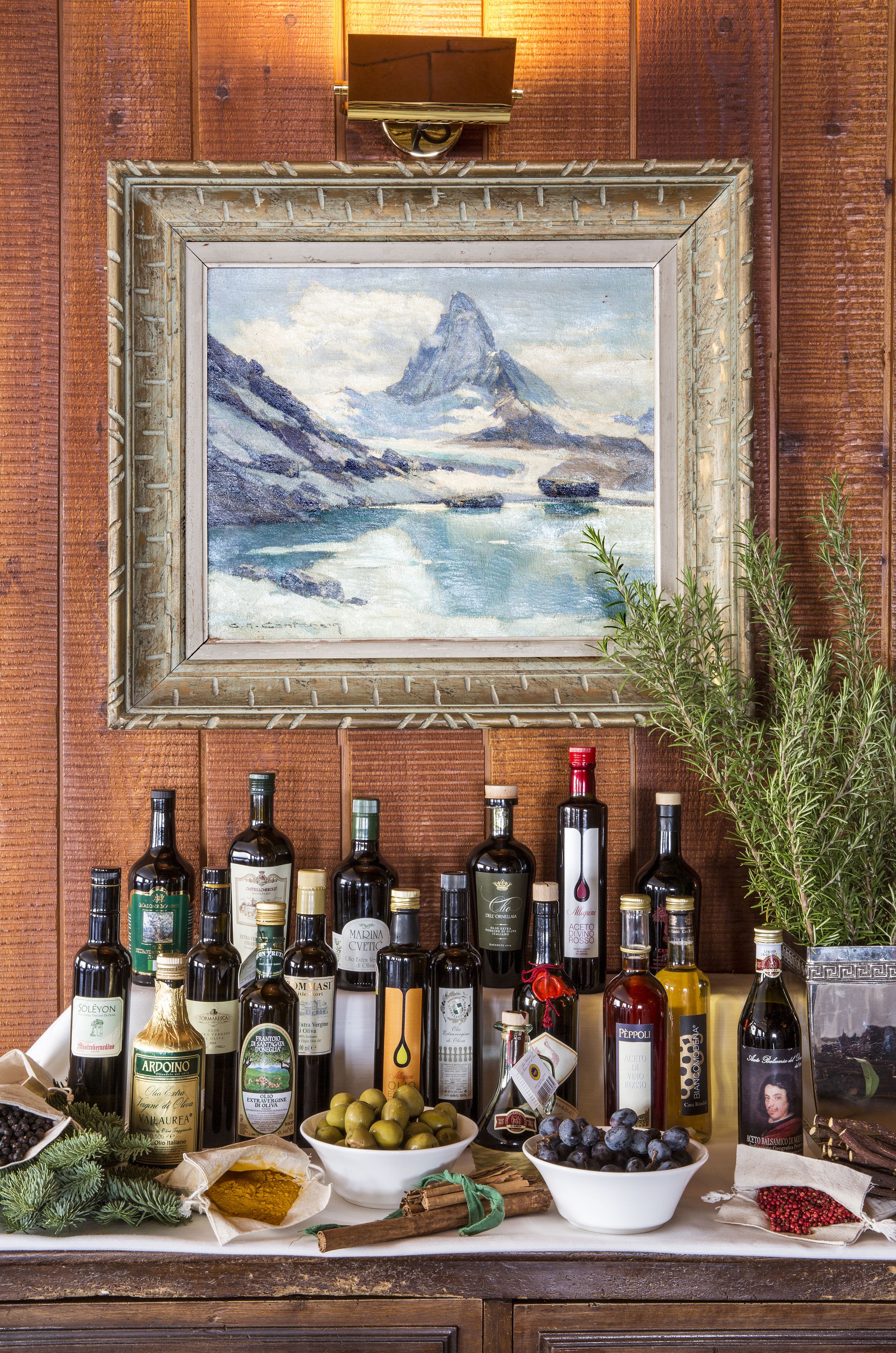 A wooden wall with a framed painting of a mountain landscape, a variety of bottles, and bowls of olives and blueberries.