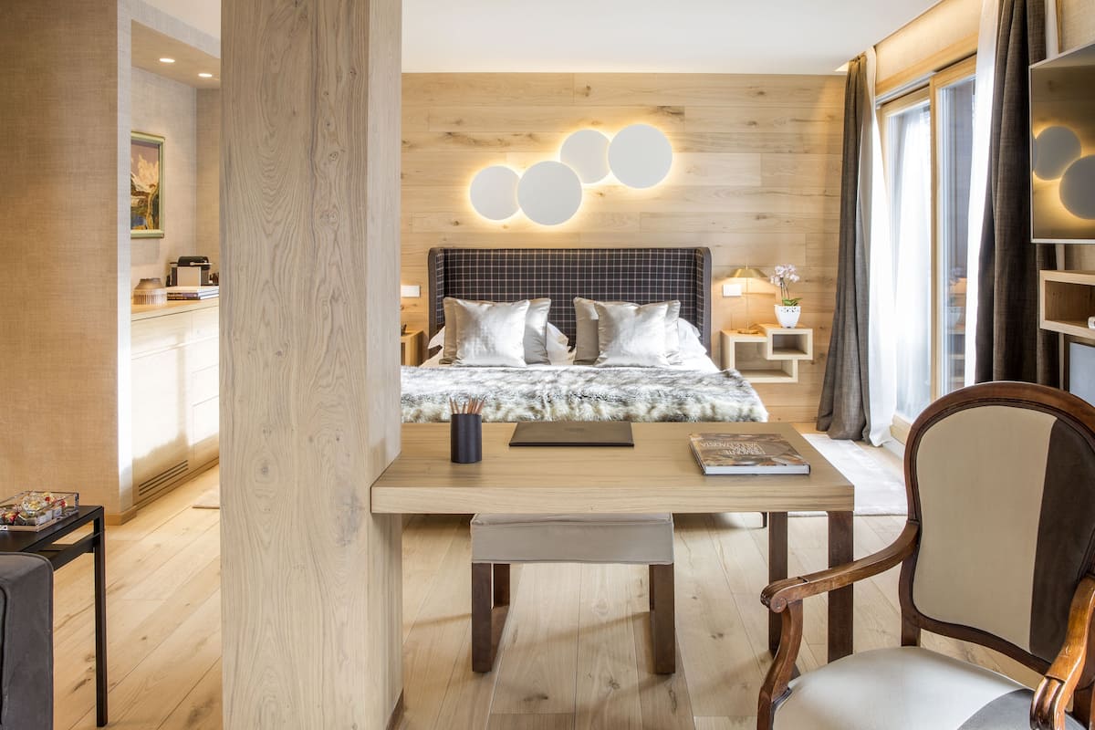 Un dormitorio moderno con cabecera de madera, una mesita de noche con lámpara y vistas a un paisaje urbano a través de amplios ventanales.