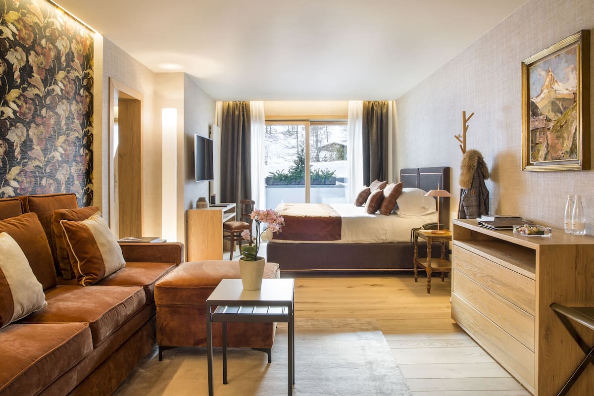 Una habitación de hotel moderna con sofá, una mesita, una cama con vistas, un aparador de madera y un cuadro en la pared.