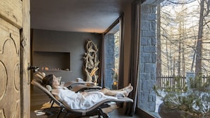 A spa room with two reclining chairs, a fireplace, and a view of trees through large windows. - Hermitage Hotel (Valtournenche)