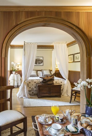 A bedroom with a canopy bed, a bedside table with a lamp, a chair, and a breakfast setup with plates, cups, and a glass of orange juice. - Hermitage Hotel (Valtournenche)