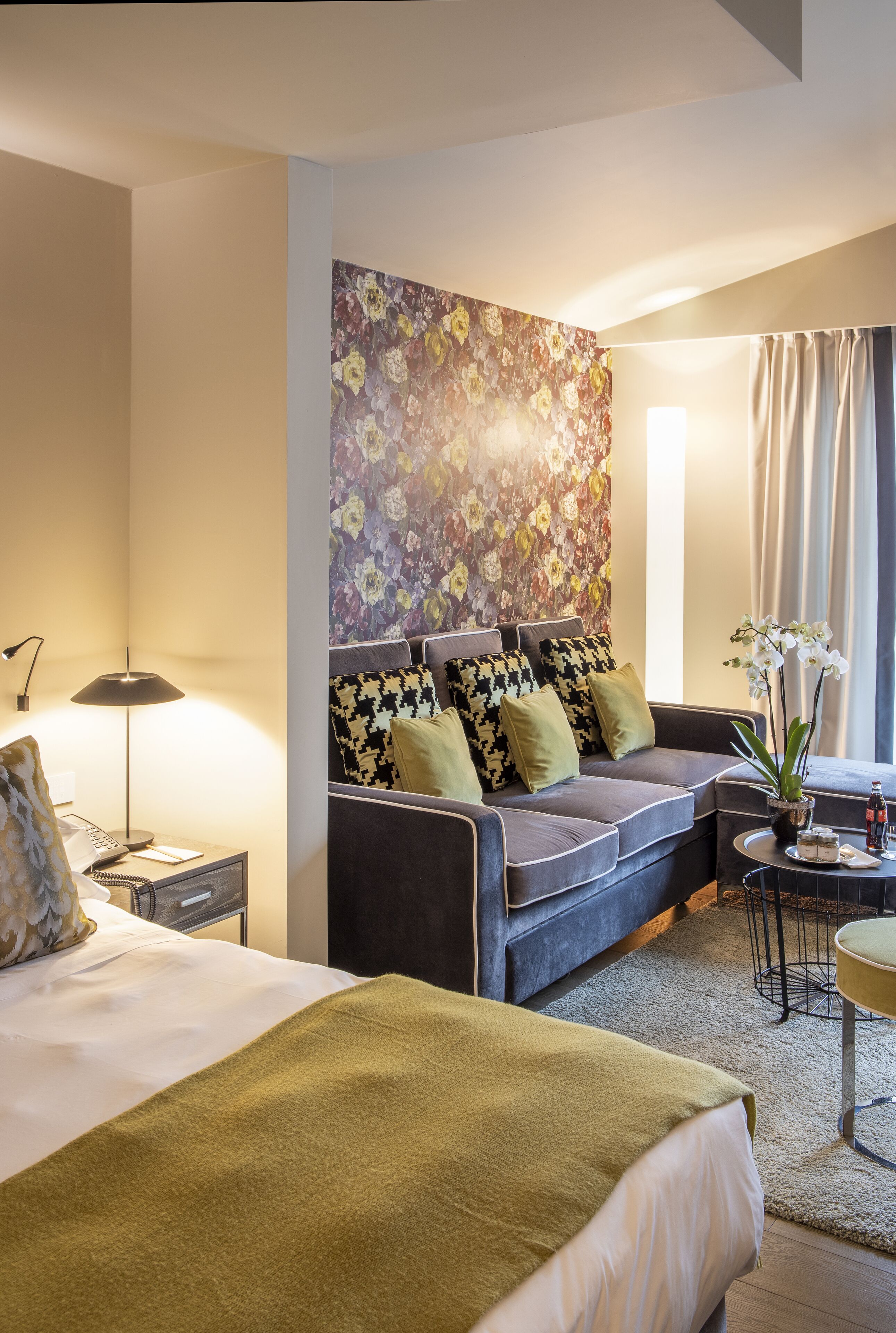 A hotel room with a bed, a sofa, a nightstand, a lamp, a floral wall mural, and a window with curtains.