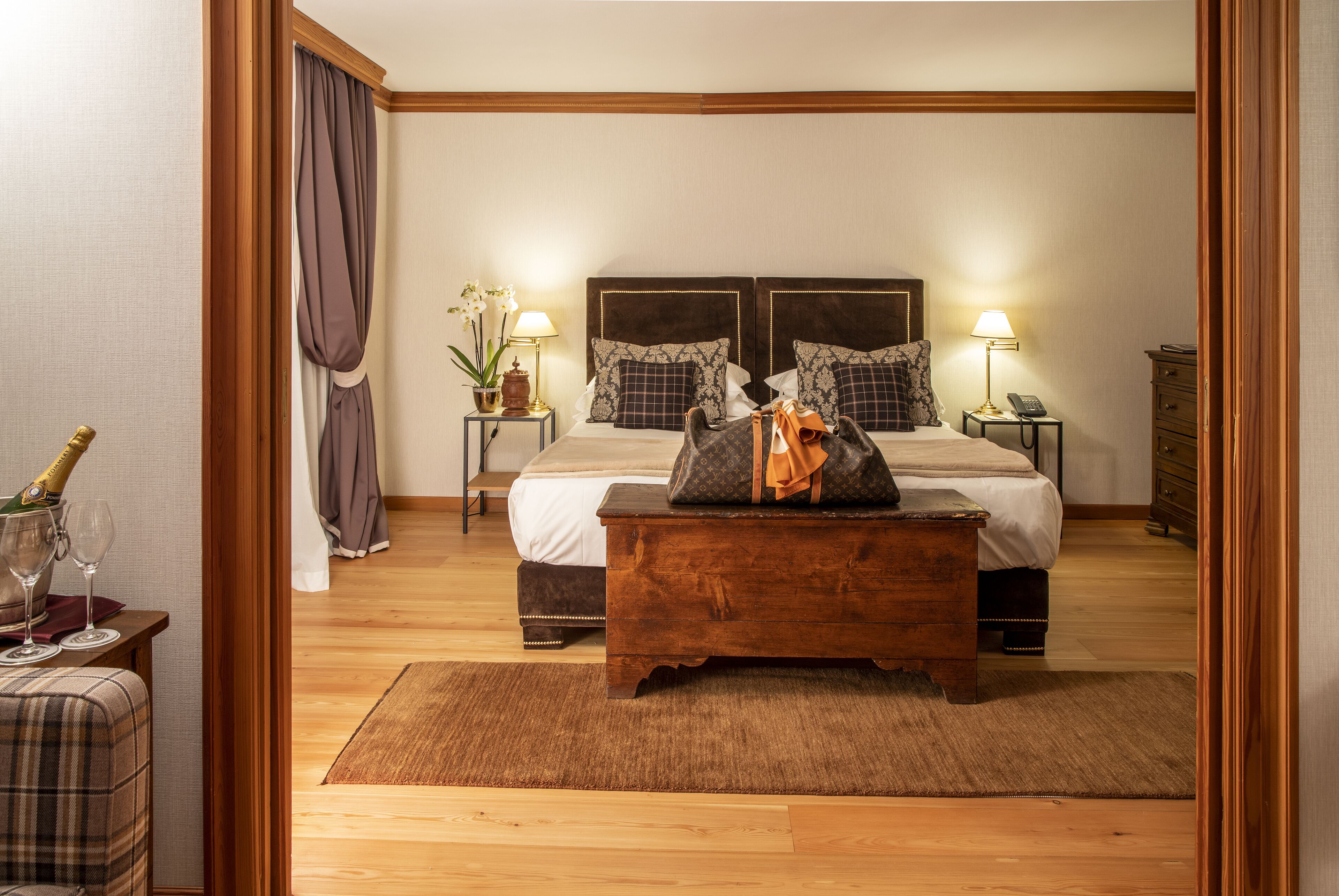 A hotel bedroom with a large bed, two bedside tables with lamps, a wooden bench, and a bottle of champagne.