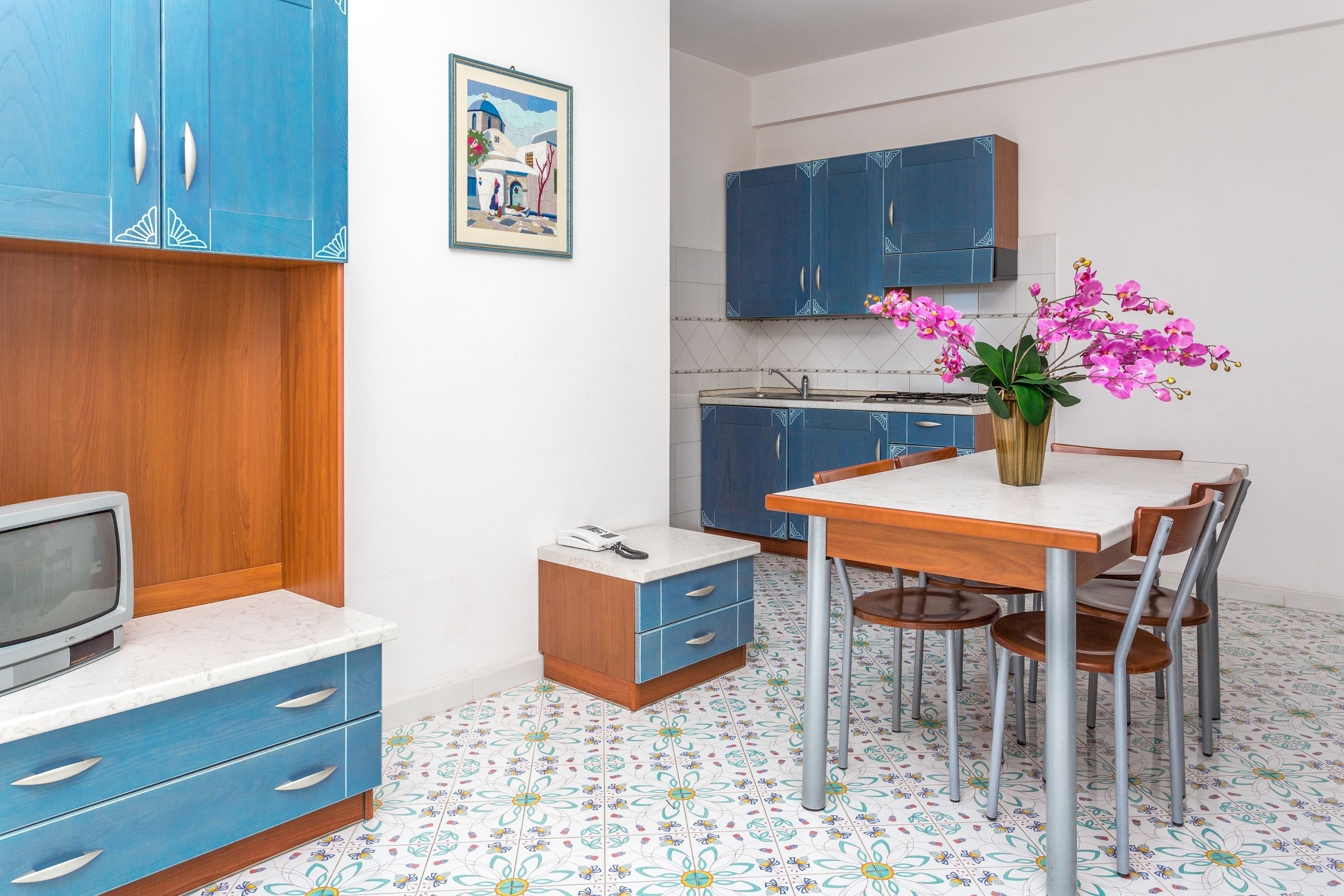A compact kitchen with blue cabinets, a dining table with chairs, a small TV, and a vase with flowers.