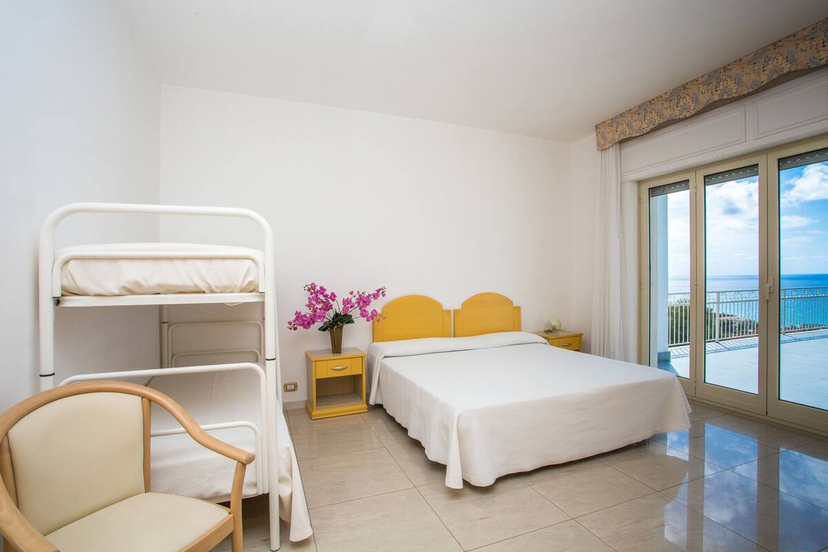 Une chambre d’hôtel avec un lit superposé, un lit simple, une chaise en bois, une table de chevet ornée d’un vase de fleurs et un balcon avec vue sur la mer.