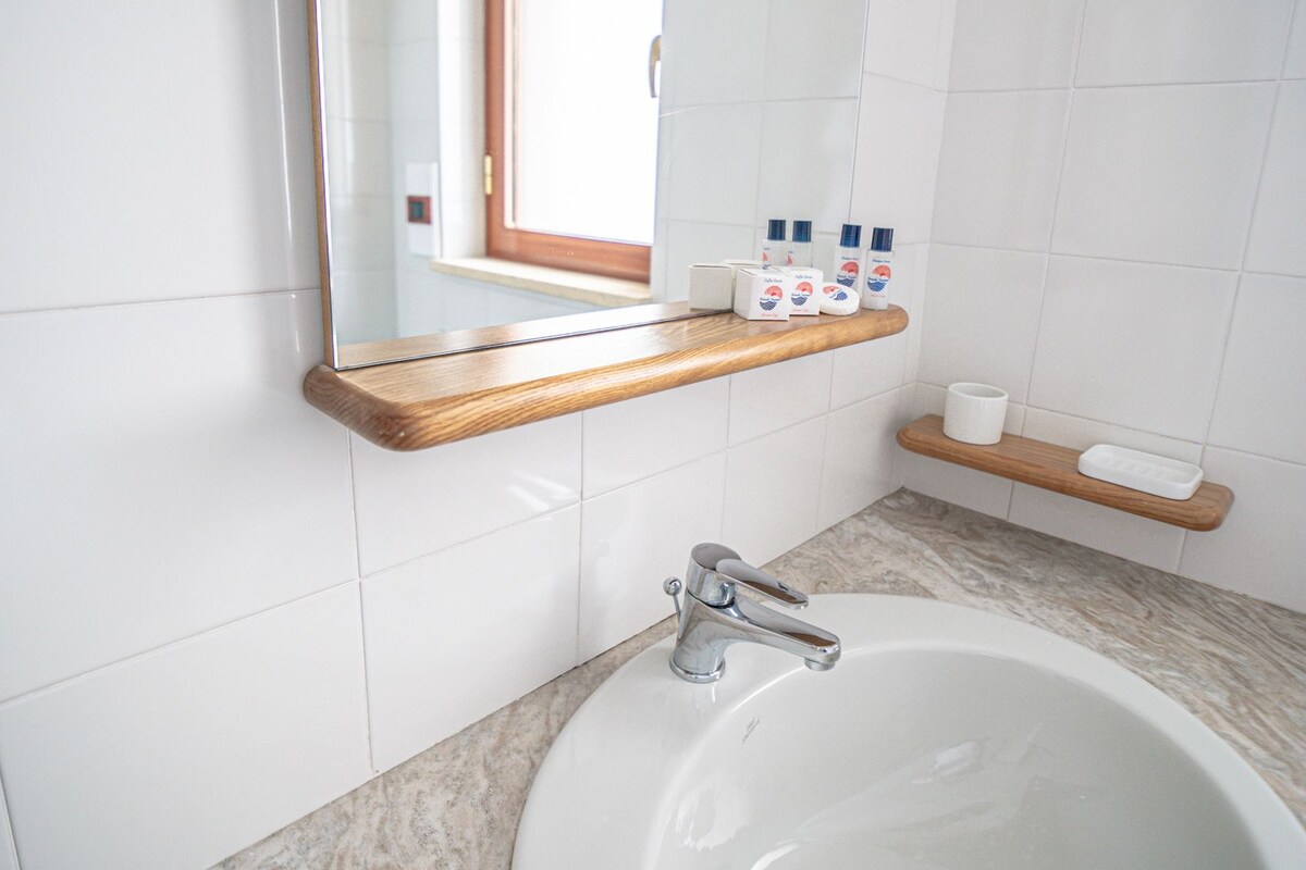 Une salle de bain avec un lavabo blanc, un miroir, une étagère en bois où sont rangés les articles de toilette et une fenêtre.