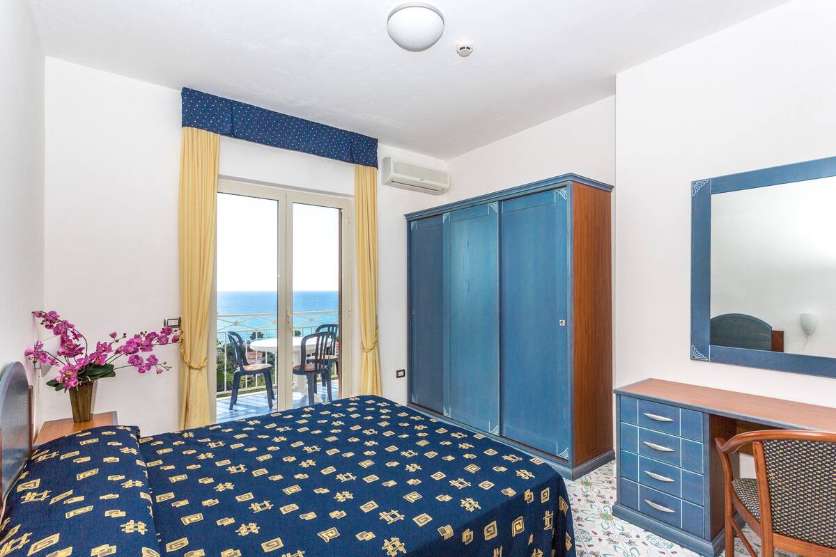 Une chambre d’hôtel avec un lit, une armoire, un bureau et un miroir. La fenêtre offre une vue sur la mer.