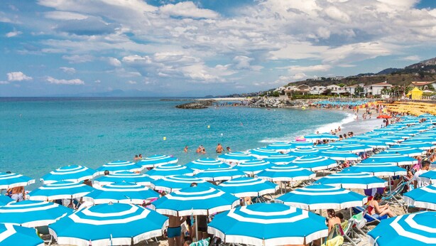 A beach with numerous blue umbrellas and people enjoying the water.