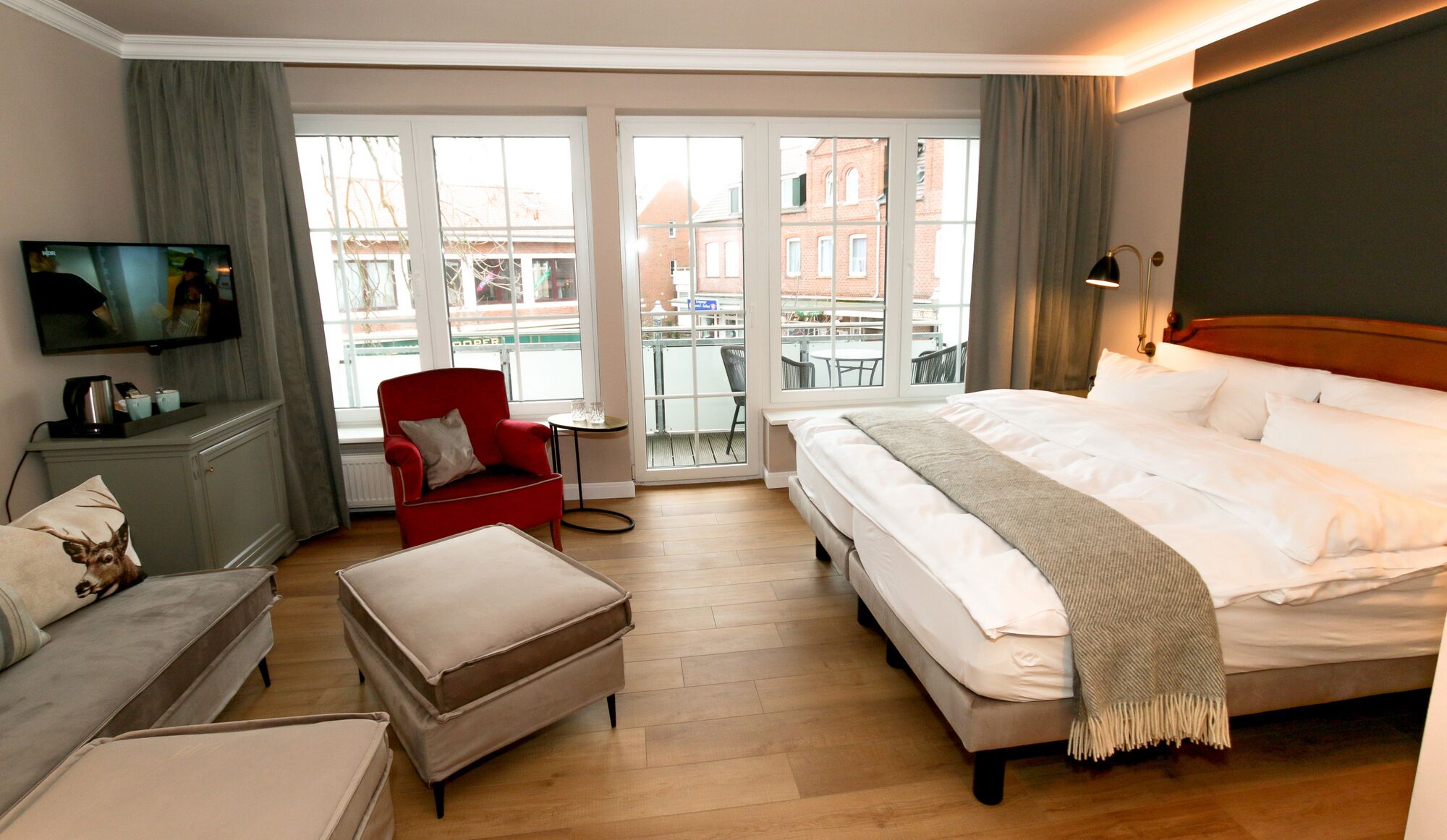 Camera d'albergo con un letto grande, una poltrona rossa, un divano, una televisione e un balcone con vista sugli edifici.
