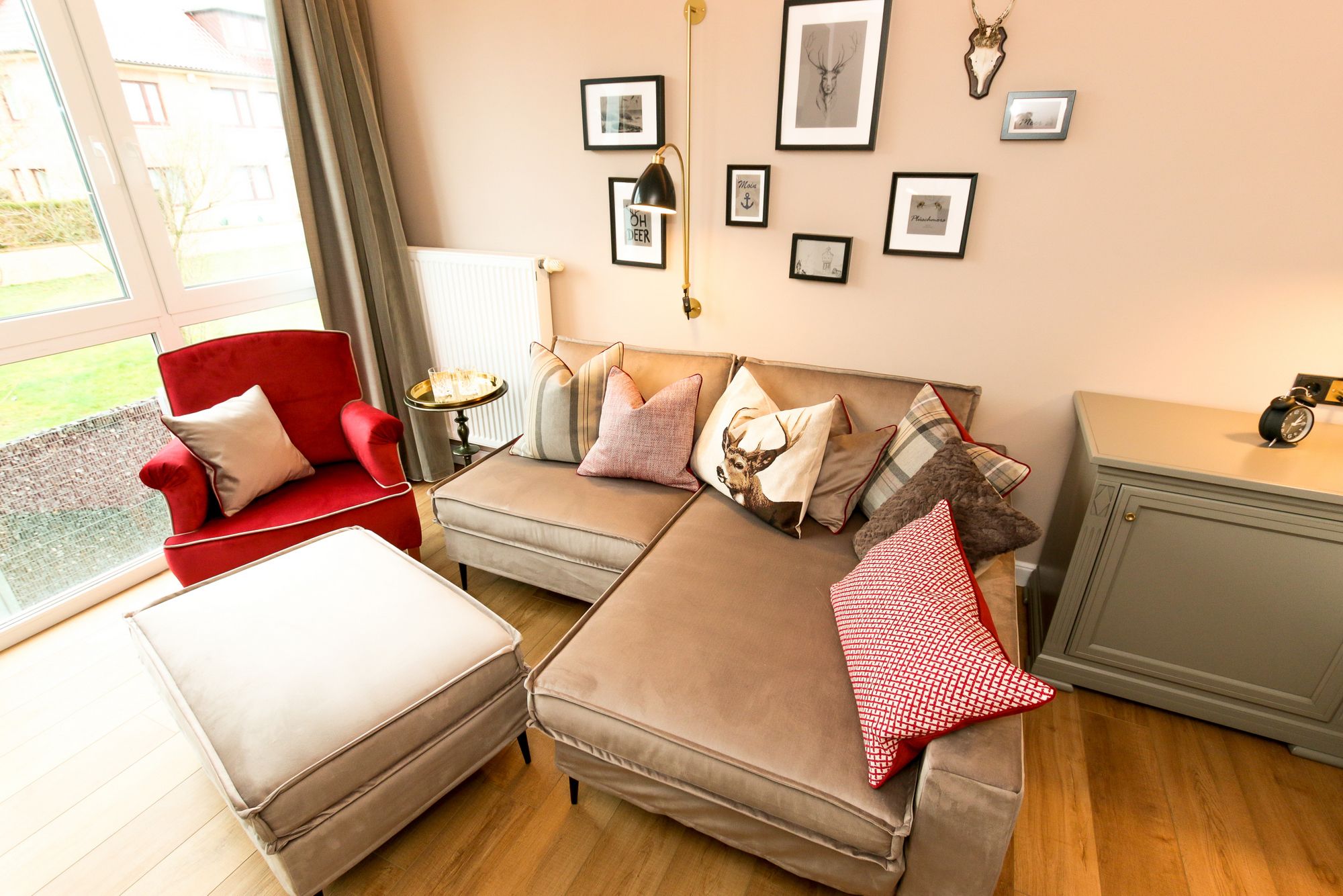 A cozy living room with a red armchair, a beige sofa, and a small table with a lamp. There are framed pictures on the wall and a window with curtains.