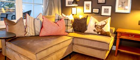 A cozy living room with a sectional sofa adorned with various patterned pillows, a small wooden side table, and framed artwork on the wall.