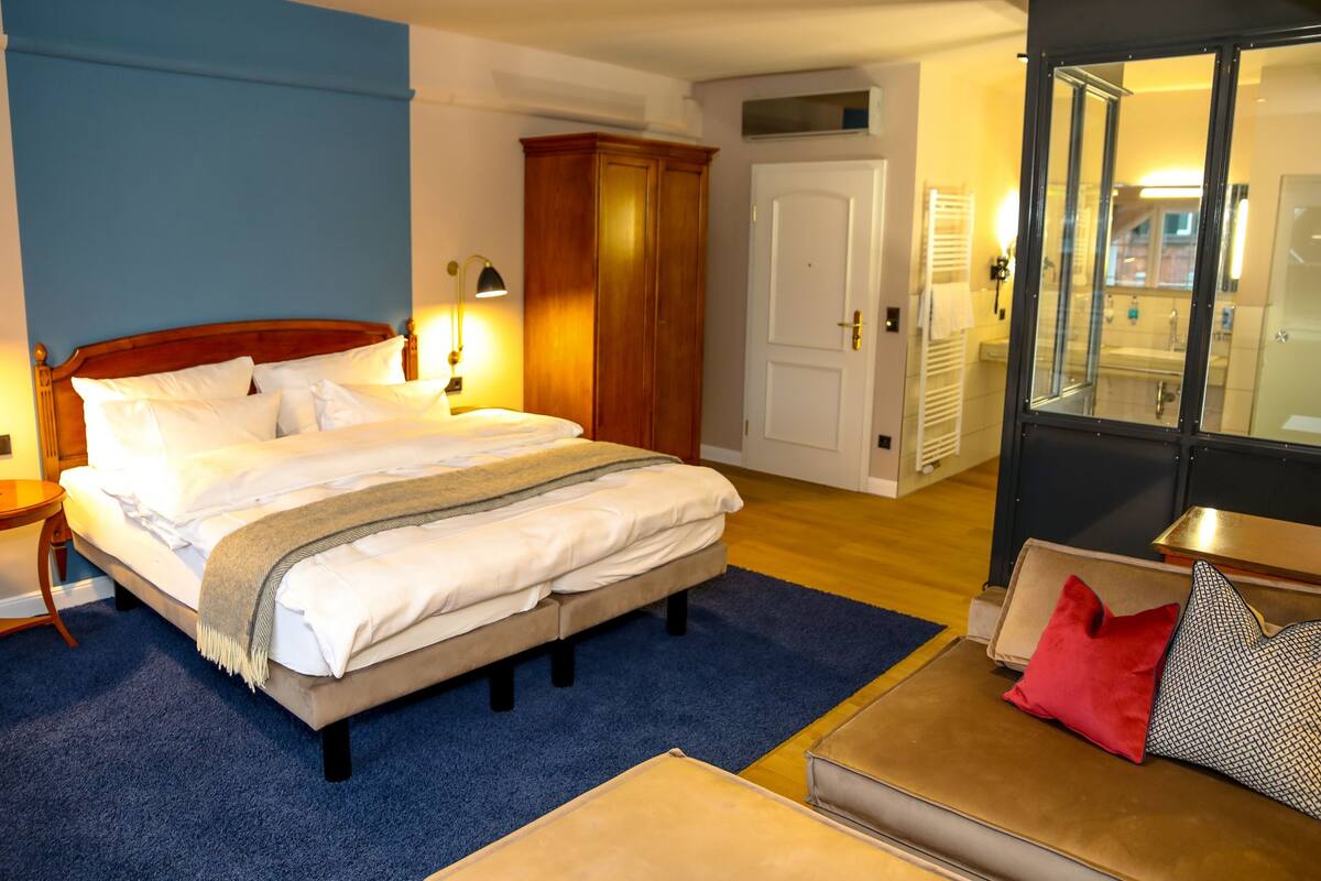 Une chambre d’hôtel avec un grand lit, des lampes de chevet, une armoire en bois, une petite table avec une horloge et une vue sur la salle de bain à travers une porte en verre.