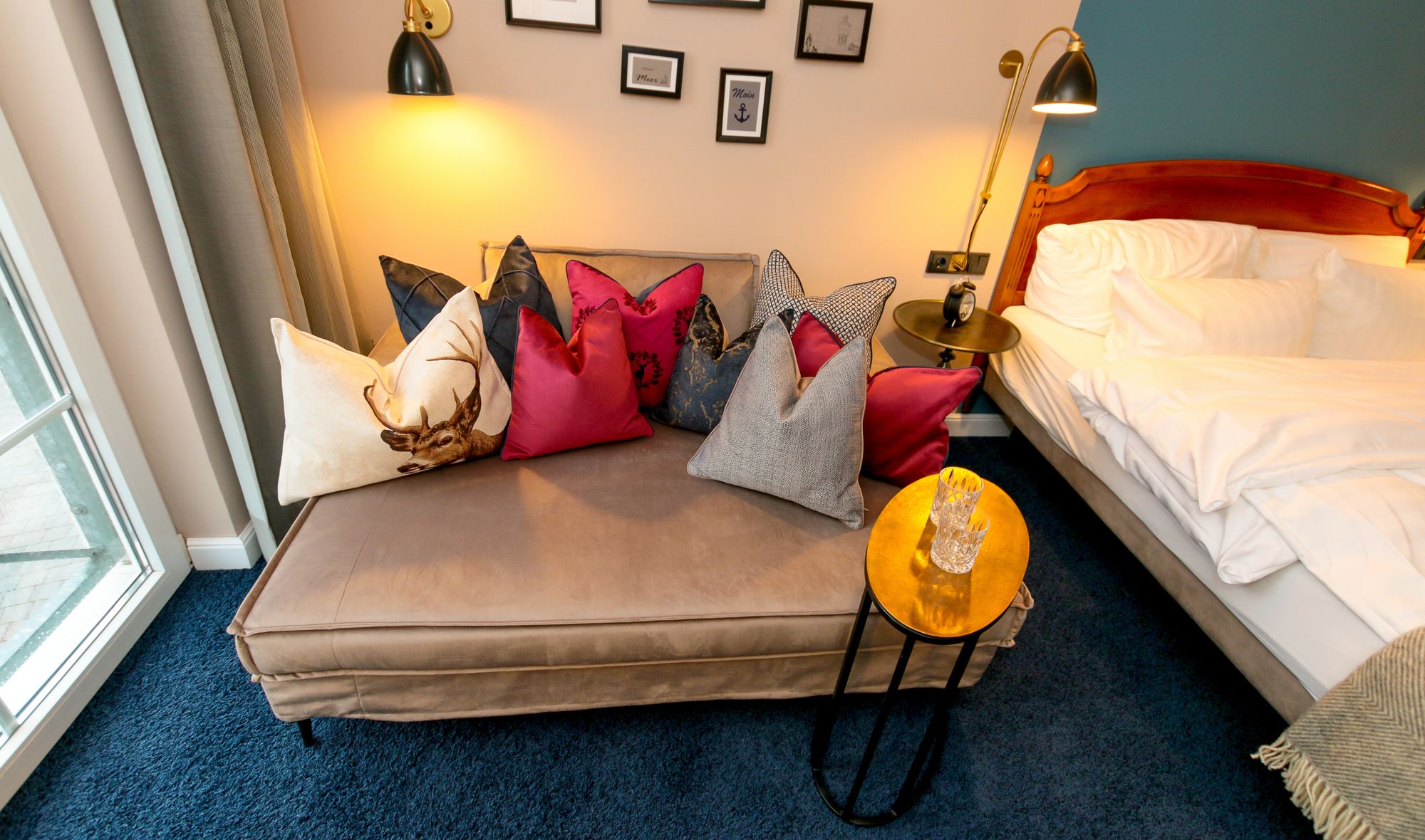 A cozy hotel room with a bed, a sofa with colorful cushions, a small round table with a glass, and a wall with framed pictures.