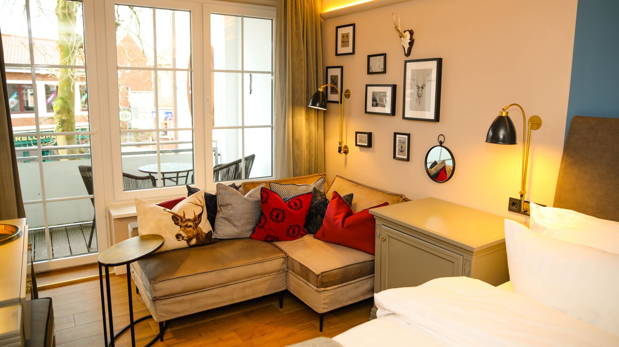 A cozy room with a sofa, a small table, a bedside table, and a bed. There are framed pictures on the wall and a window with curtains.