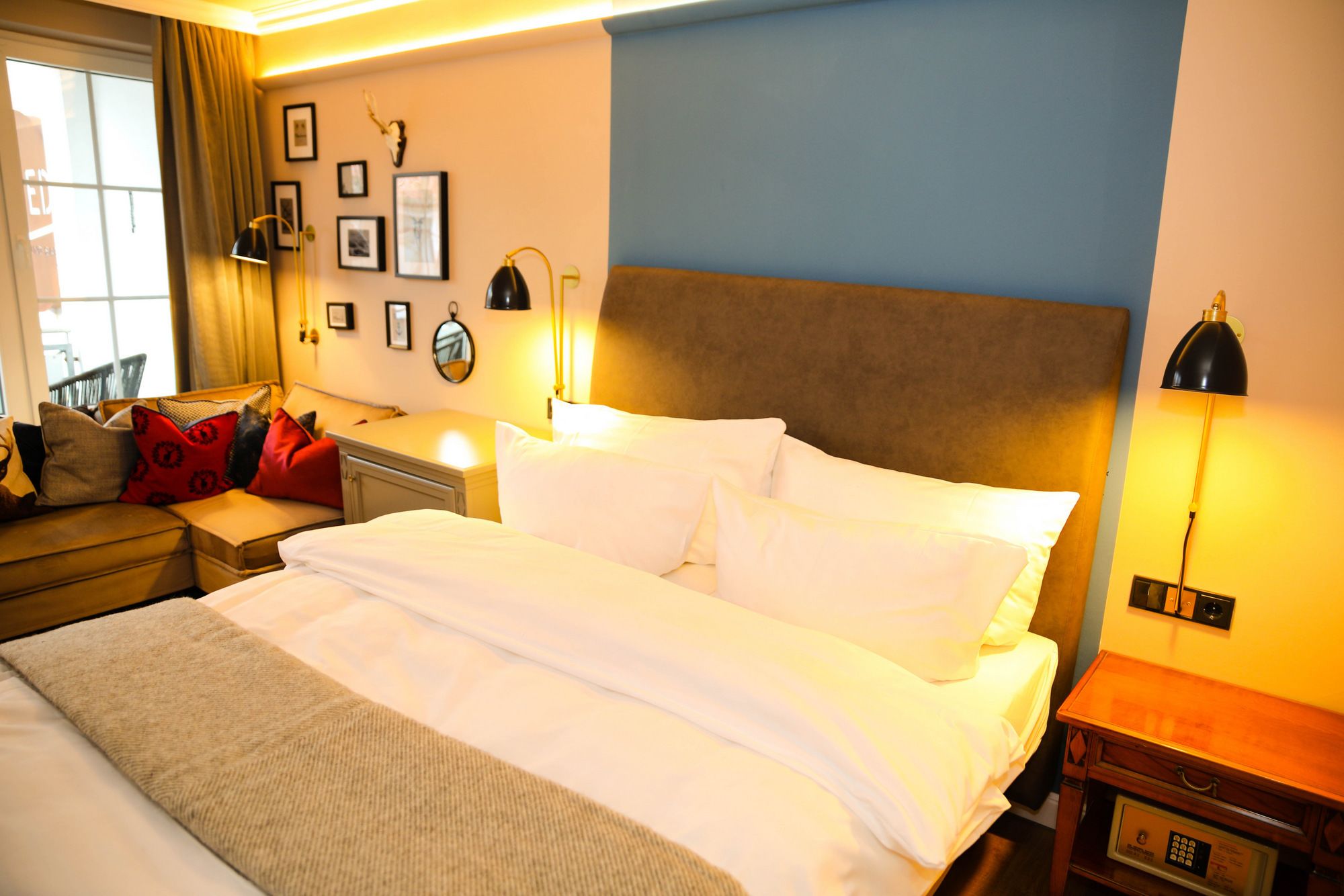 A hotel room with a large bed, a bedside table with a lamp, a nightstand, a sofa with cushions, and a wall with framed pictures and a mirror.