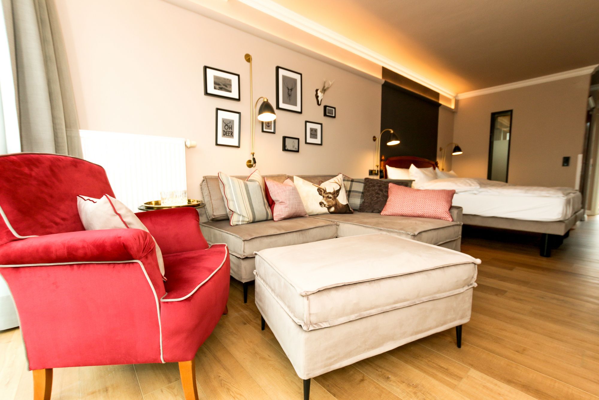 A modern hotel room with a red armchair, a grey sofa, and a white ottoman. There is a bed with white bedding, a nightstand with a lamp, and a wall adorned with framed pictures and a deer skull.