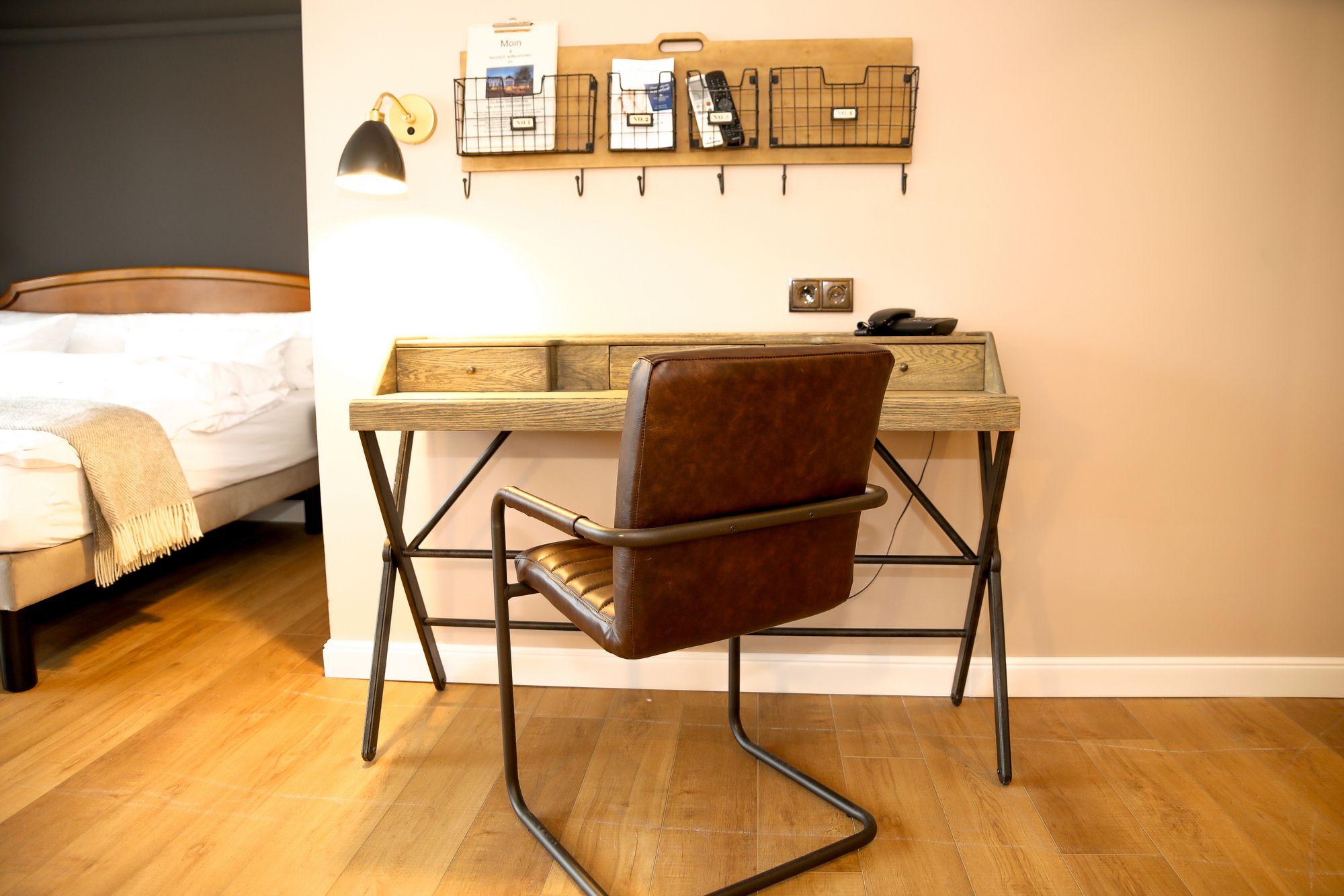 Una camera da letto con una scrivania in legno, una poltrona in pelle marrone, un letto con copriletto bianco e una mensola a muro con libri e una lampada.