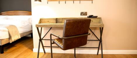 Una camera da letto con una scrivania in legno, una poltrona in pelle marrone, un letto con copriletto bianco e una mensola a muro con libri e una lampada.