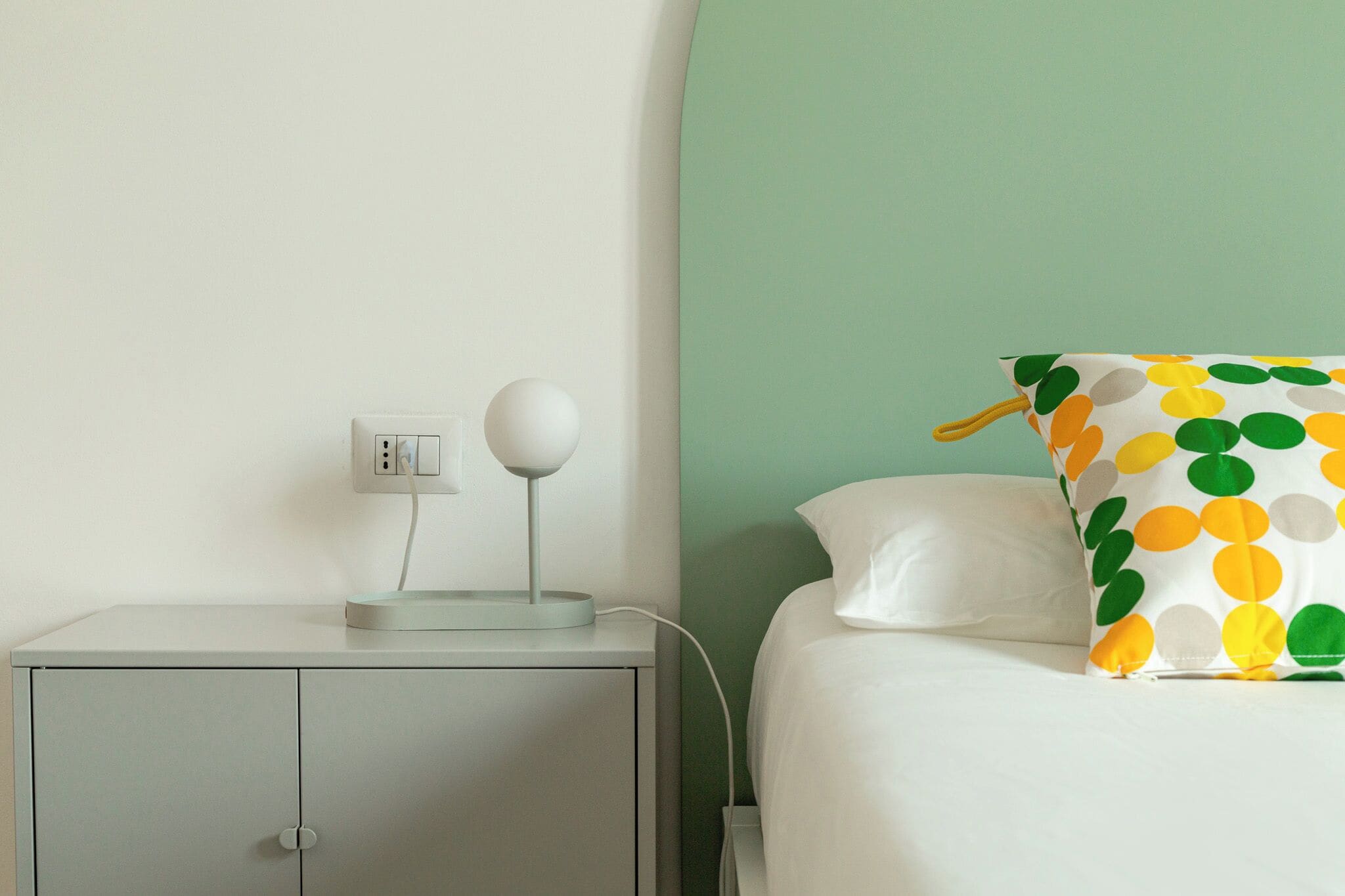 Un dormitorio ordenado con una mesita de noche, una lámpara y una cama con un cojín de lunares.