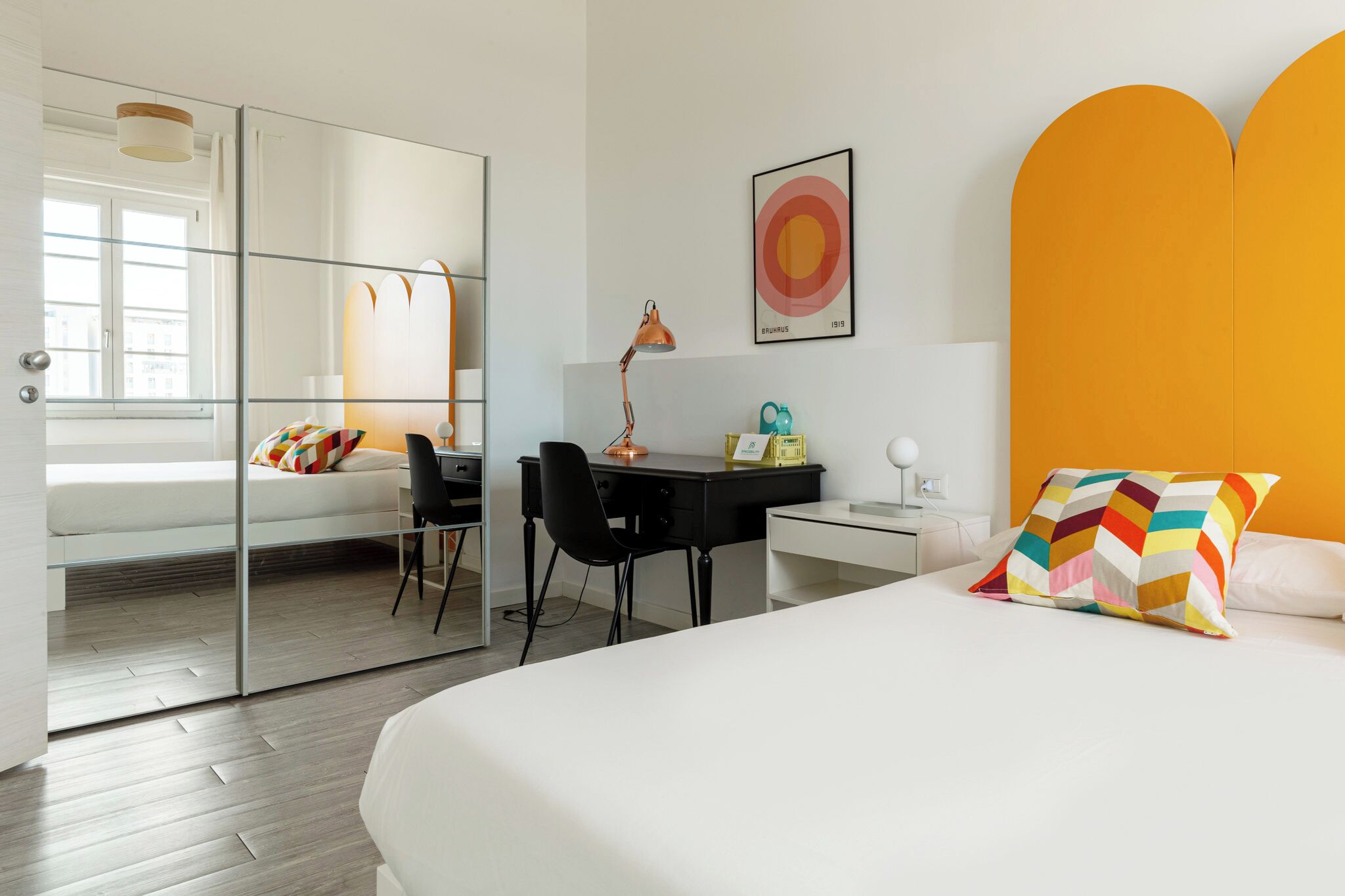 Ein modernes Hotelzimmer mit Bett, Schreibtisch, Stuhl, Lampe und einem farbenfrohen Kissen.