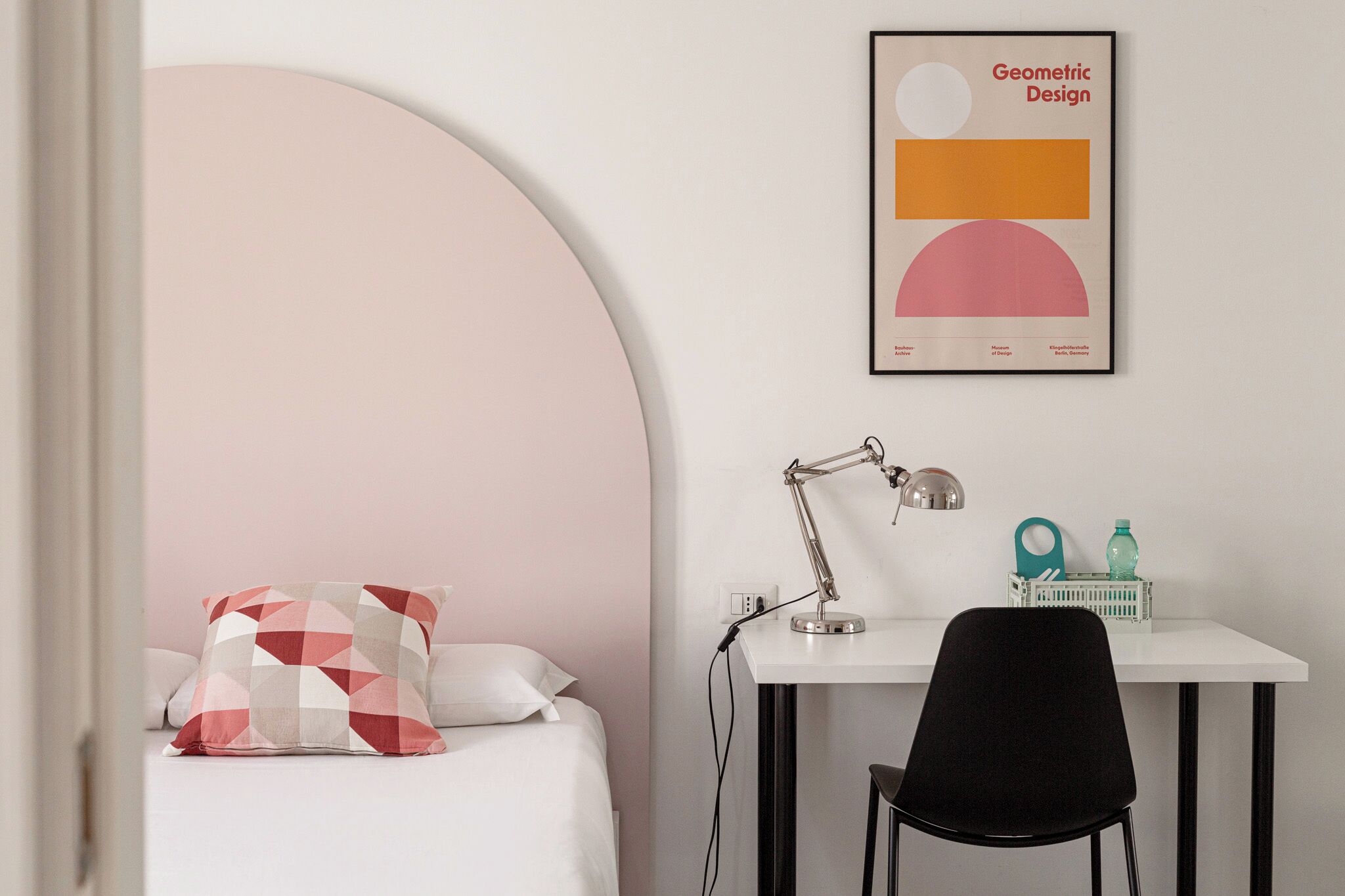 Un dormitorio con una cama, un escritorio con una silla, una lámpara y un póster con un diseño geométrico enmarcado en la pared.