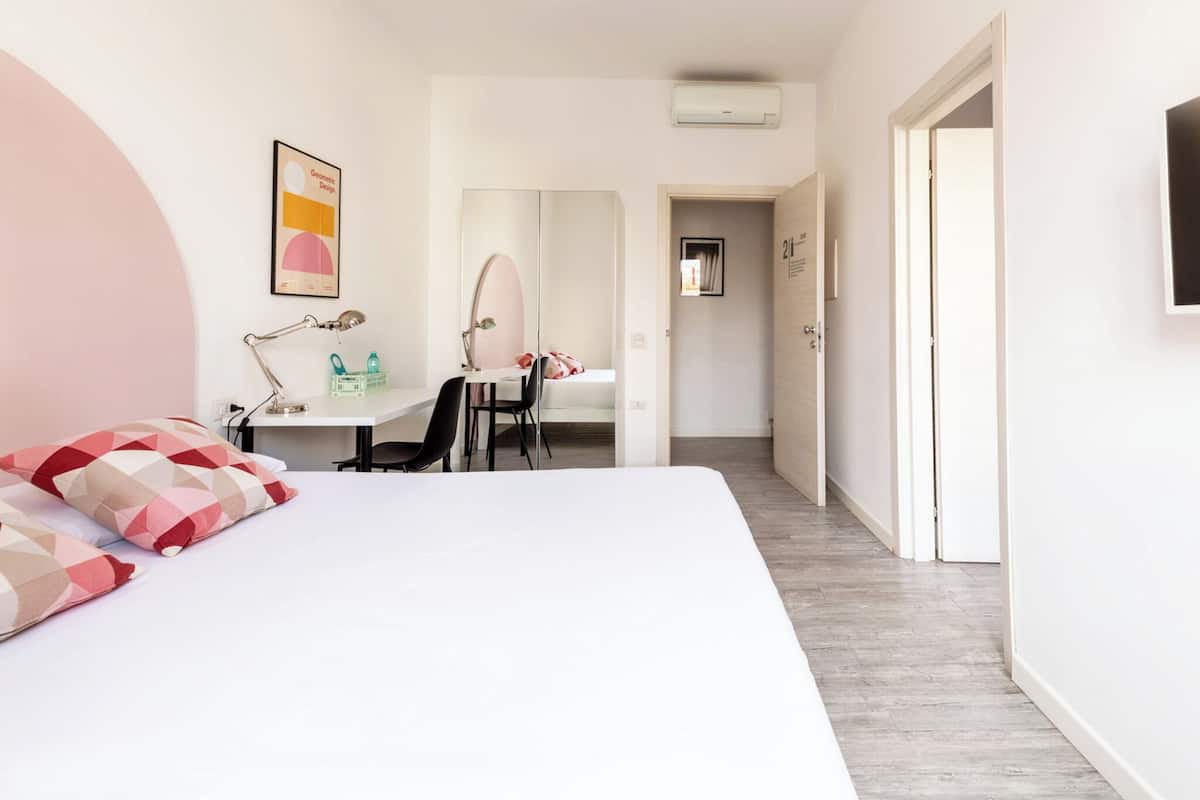 A modern bedroom with a large bed, a desk with a chair, a mirror, and a door leading to another room.