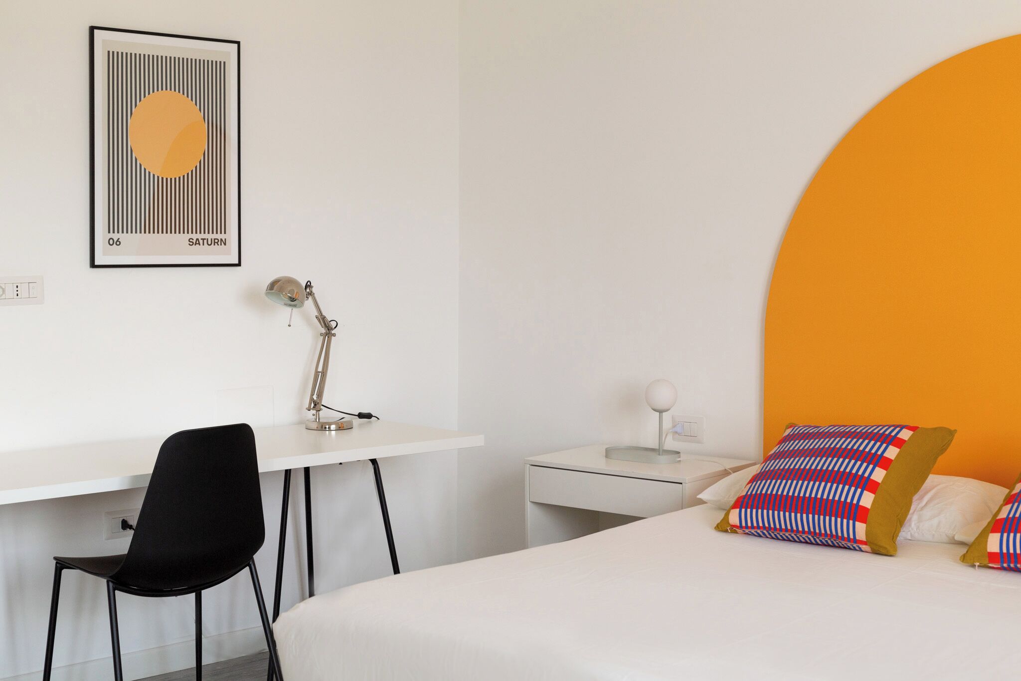 Une chambre d’hôtel avec un bureau blanc, une chaise noire, un lit agrémenté d’oreillers colorés et un tableau représentant Saturne accroché au mur.