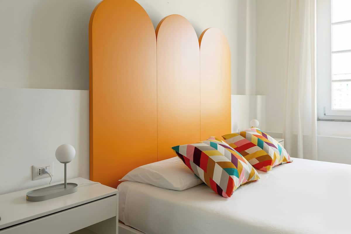 A modern bedroom with a white bed, a colorful geometric pillow, a white bedside table with a lamp, and a plain orange headboard.