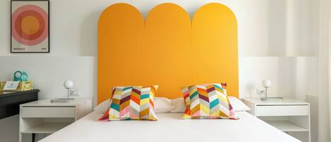 A modern hotel room with a white bed, colorful chevron-patterned pillows, a bright orange headboard, and a minimalist design.