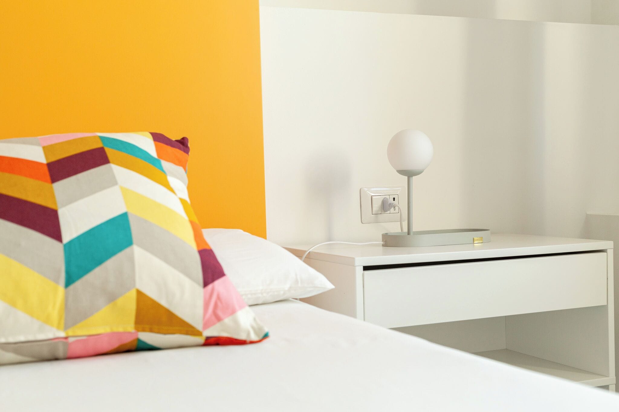 A modern hotel room with a white bed, a colorful chevron-patterned pillow, a white nightstand, and a bedside table with a lamp.