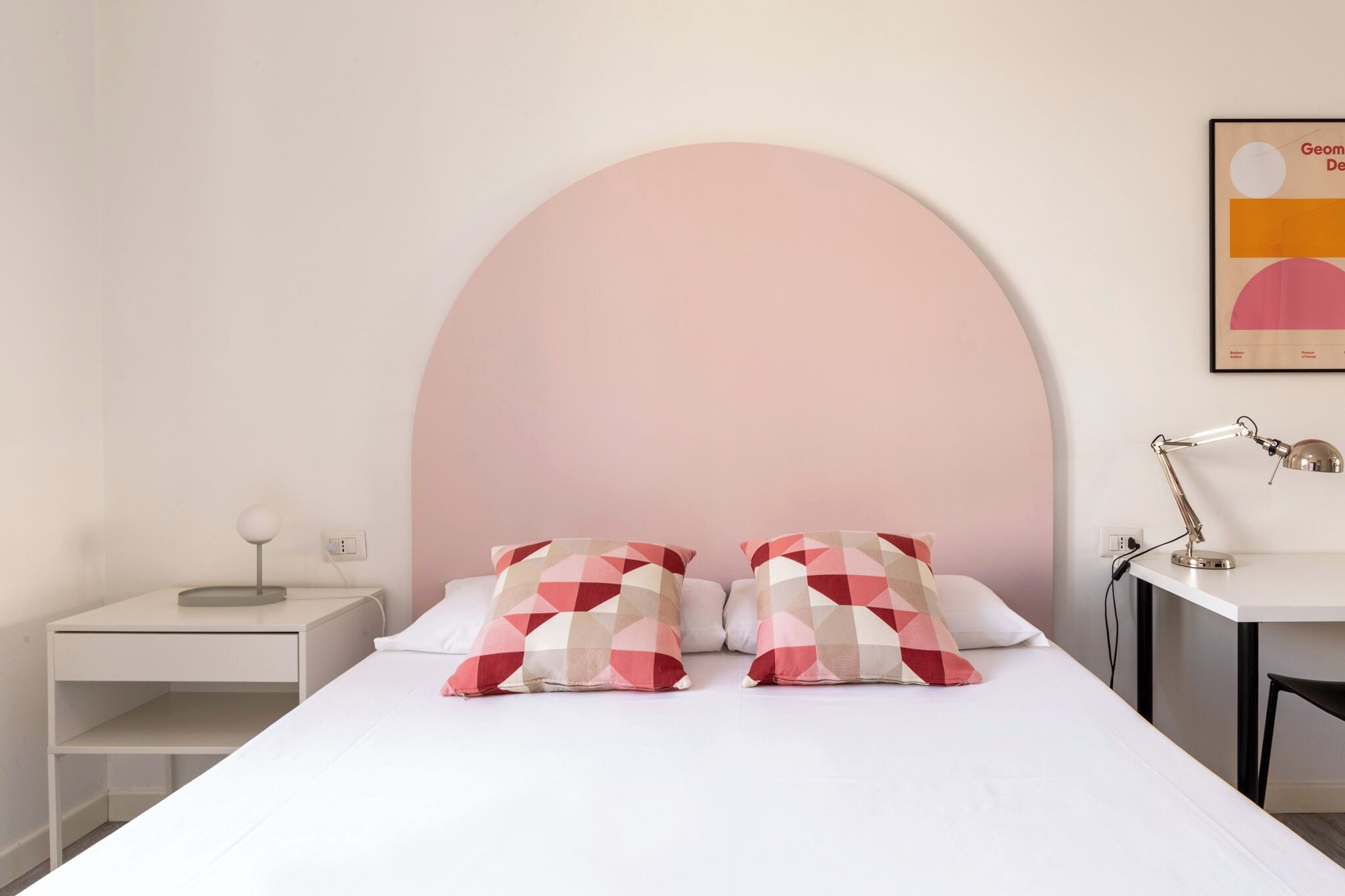 Ein Schlafzimmer mit einem Bett, zwei geometrisch gemusterten Kissen, einem Nachttisch mit Lampe und einem Schreibtisch mit Lampe.