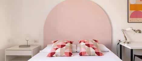 Un dormitorio con una cama, dos almohadas de diseño geométrico, una mesita de noche con lámpara y un escritorio con lámpara.