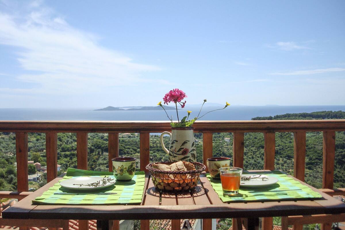 A table set with a vase of flowers, a bowl of nuts, a plate, a cup, and a glass of drink on a balcony with a scenic view.