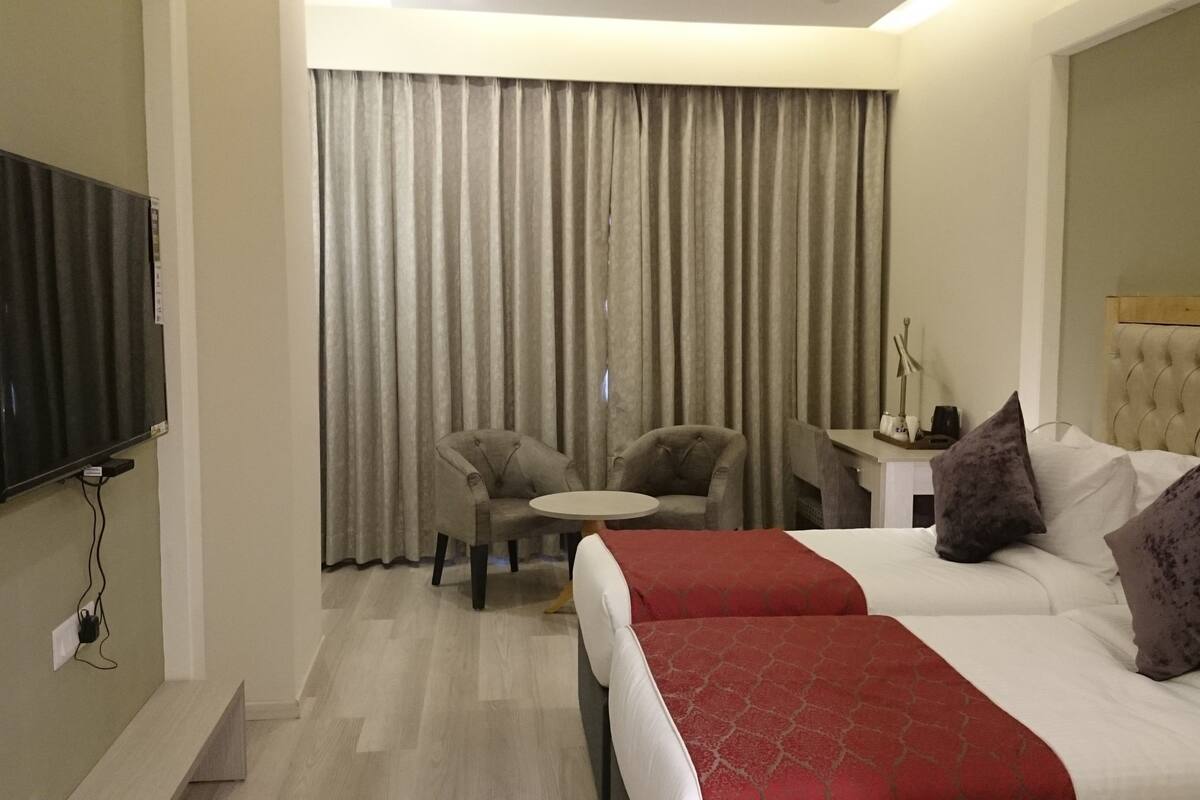 Quarto de hotel com cama, duas poltronas, uma mesinha redonda, televisão fixada na parede e janela com cortinas.