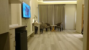 A hotel room with a flat-screen TV mounted on the wall, a small desk with a lamp, two chairs, and a wooden floor. - Mango Hotels Naveen (Davanagere)