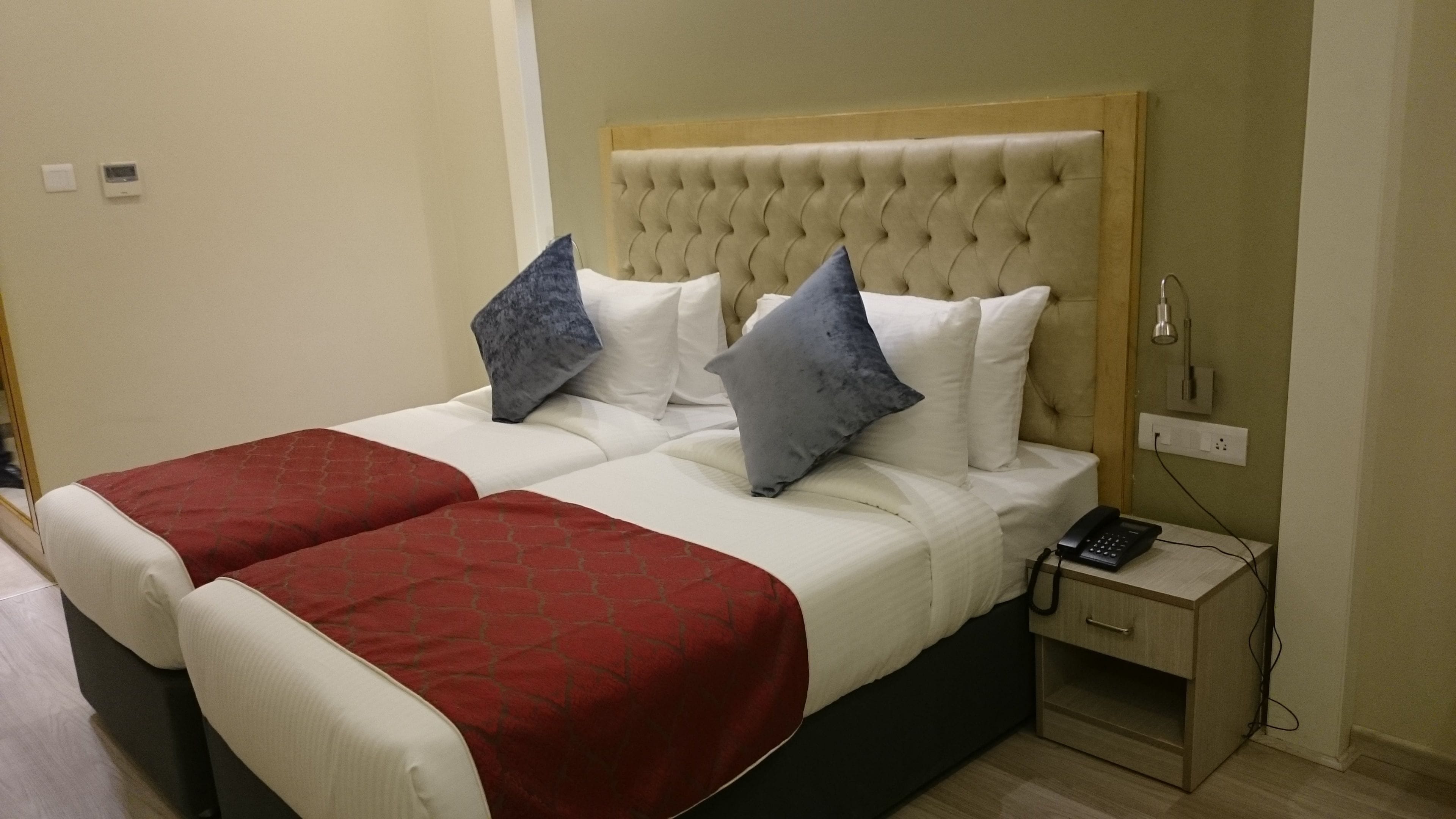 A double bed with a red and white bedspread, two grey pillows, and a wooden headboard. There is a bedside table with a phone and a lamp.