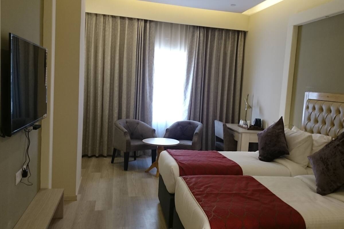 Quarto de hotel com duas camas, televisão, escrivaninha com cadeira e janela com cortinas.
