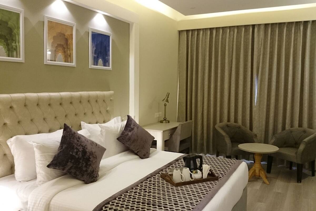 Quarto de hotel com cama de cabeceira estofada, duas poltronas, uma mesa pequena e uma janela grande com cortinas.