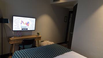 A hotel room with a bed, a desk with a computer, a chair, and a TV displaying a welcome screen in multiple languages.