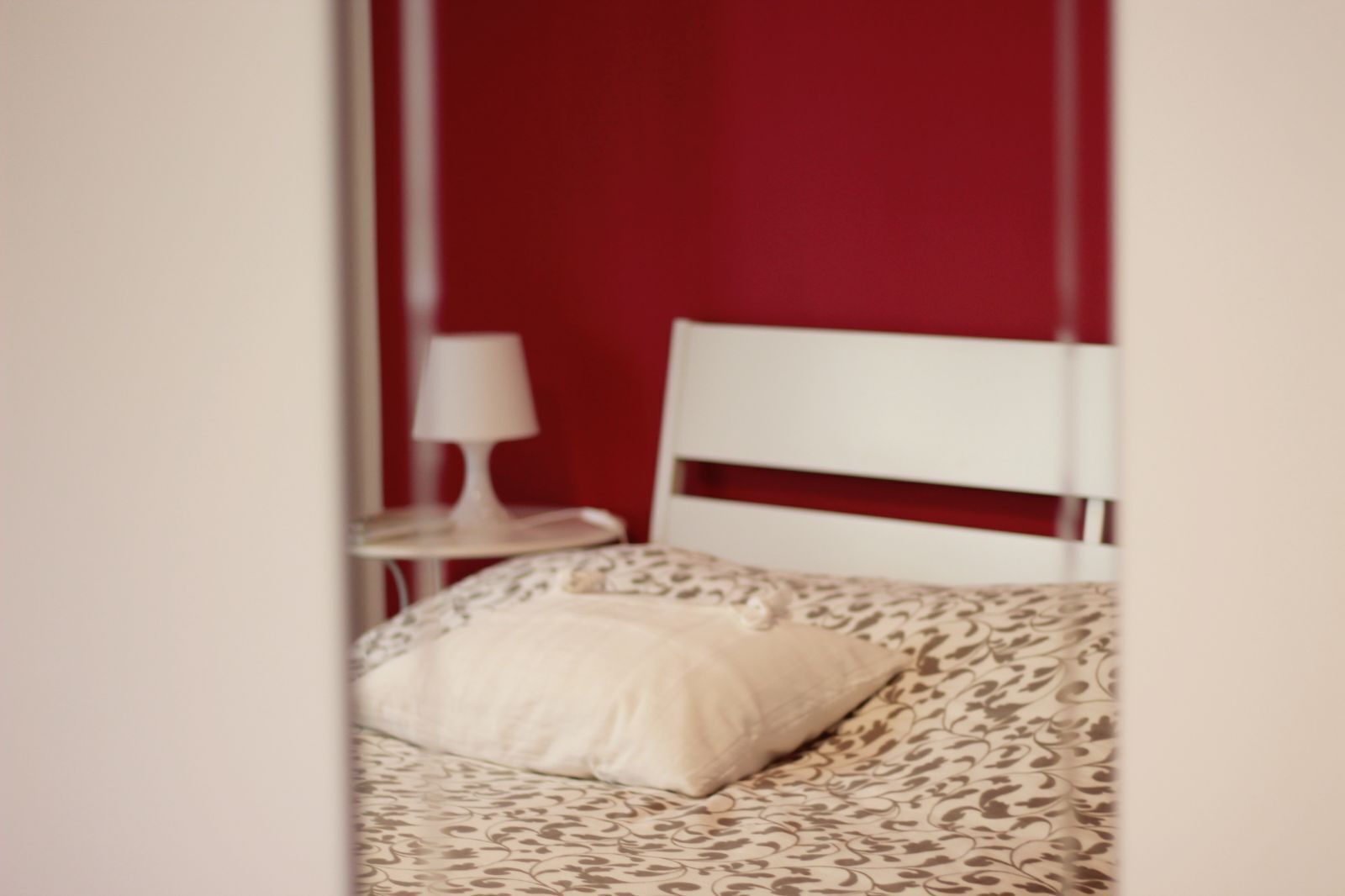 Una cama con una colcha estampada y una sola almohada, una mesita de noche blanca con una lámpara y una pared roja.