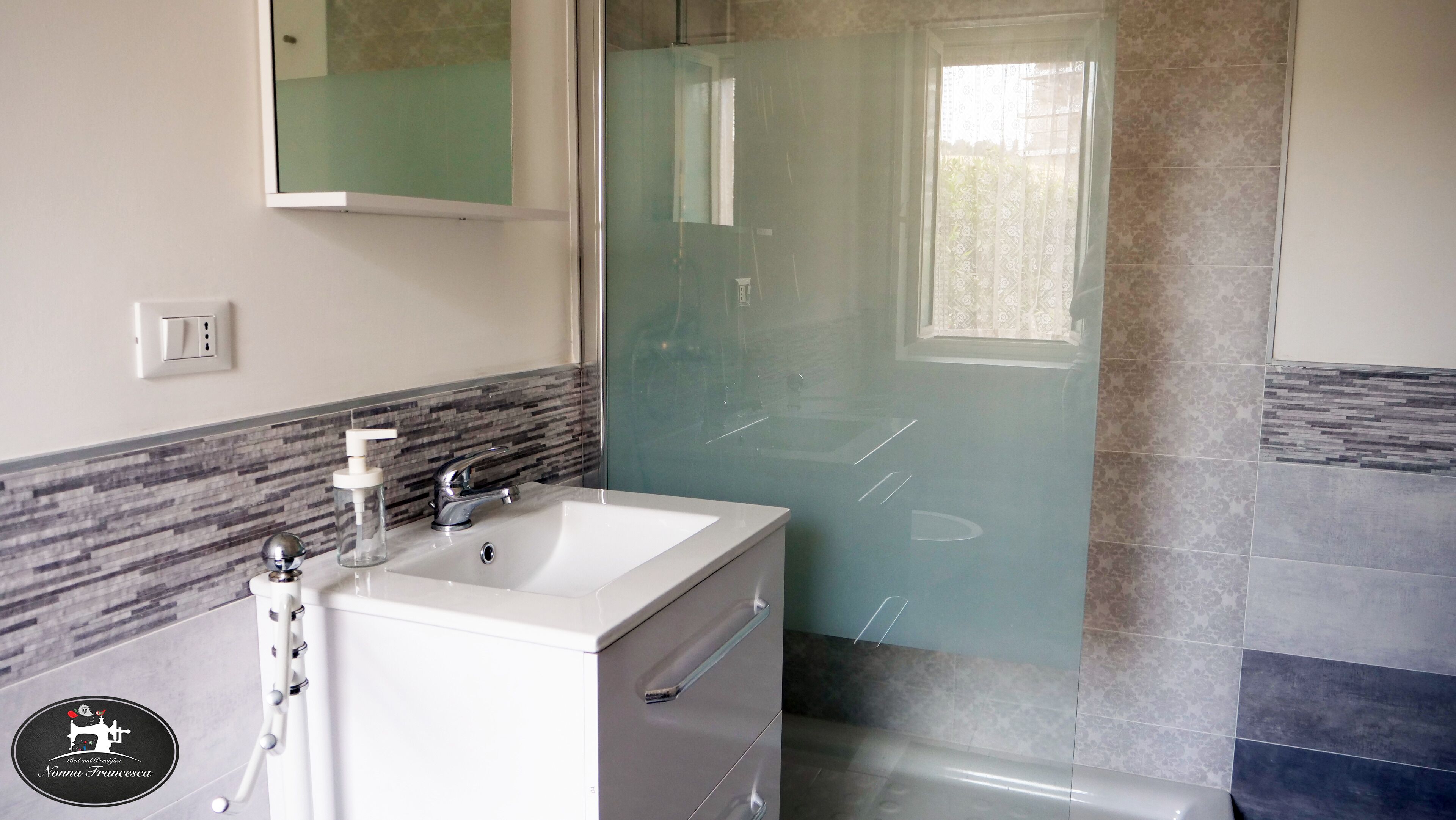 Un baño con lavamanos blanco, espejo, cabina de ducha de vidrio y pared con azulejos.